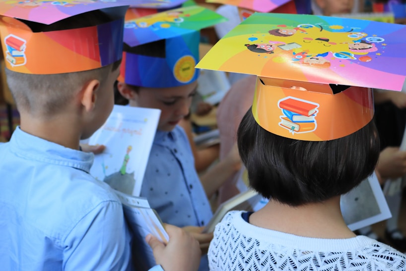 Children wearing colorful paper hats with educational-themed designs are viewing illustrated books. The hats are decorated with images of books and cartoon faces, suggesting a celebration or educational event.