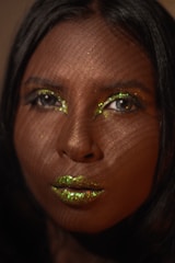 A close-up portrait of a person's face with shimmering green glitter makeup on the eyelids and lips. The lighting creates a patterned shadow effect across the face, adding texture and depth.