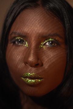 A close-up portrait of a person's face with shimmering green glitter makeup on the eyelids and lips. The lighting creates a patterned shadow effect across the face, adding texture and depth.