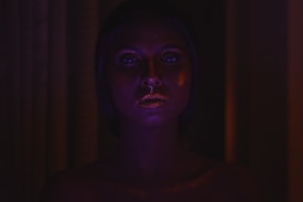 A person with glitter on their face is dimly lit in a dark environment, highlighting their facial features. The lighting casts a mysterious and ethereal glow, emphasizing the shine on their face.