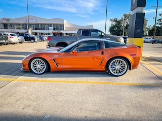 A sleek orange sports car is parked in a lot, surrounded by other vehicles. It has large, shiny wheels and a streamlined body. The background shows several commercial buildings, including a nail salon and various shops.