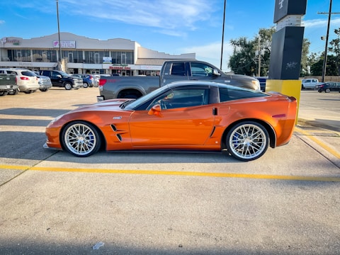 A sleek orange sports car is parked in a lot, surrounded by other vehicles. It has large, shiny wheels and a streamlined body. The background shows several commercial buildings, including a nail salon and various shops.