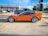A sleek orange sports car is parked in a lot, surrounded by other vehicles. It has large, shiny wheels and a streamlined body. The background shows several commercial buildings, including a nail salon and various shops.
