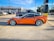 A sleek orange sports car is parked in a lot, surrounded by other vehicles. It has large, shiny wheels and a streamlined body. The background shows several commercial buildings, including a nail salon and various shops.