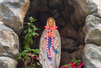 A statue of a woman in a white robe adorned with a blue sash, situated within a rocky grotto. The statue is decorated with a pink and red rosary, and there are green and red plants surrounding it.