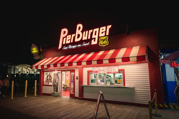 A retro-style burger joint with a red and white striped canopy, illuminated by bright neon signage. The Pier Burger logo is prominently displayed, and a Route 66 emblem adds a vintage feel. The establishment features takeout windows with visible menu signs.