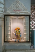 An intricately carved alcove holds a brightly adorned deity statue decorated with yellow and orange marigold flowers. The statue depicts a figure in colorful garments riding a white horse, enclosed within a glass case framed by ornate white pillars.