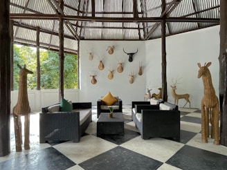 A contemporary seating area with black wicker furniture and colorful cushions arranged on a checkered black and white floor. Large wicker giraffe sculptures stand on either side, and several animal head mounts decorate the white walls. The space is open to greenery outside, with corrugated metal roofing supported by rustic wooden beams.