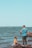 A man is standing on rocky terrain by the water, holding a fishing rod, while a child in a yellow swimsuit plays nearby, splashing in the water. The sky is clear with light blue tones, and the background shows a wide expanse of water with distant shores.