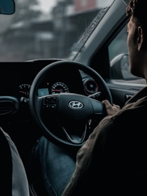 A person is seated in the driver's seat of a car, holding the steering wheel with one hand. The dashboard is visible, showing speedometer dials with faint red accents. The interior appears dimly lit, and there's a sense of focus and concentration.