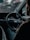 A person is seated in the driver's seat of a car, holding the steering wheel with one hand. The dashboard is visible, showing speedometer dials with faint red accents. The interior appears dimly lit, and there's a sense of focus and concentration.