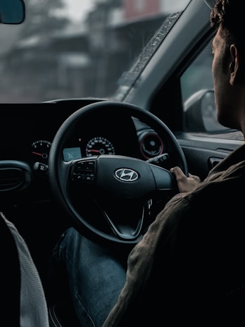 A person is seated in the driver's seat of a car, holding the steering wheel with one hand. The dashboard is visible, showing speedometer dials with faint red accents. The interior appears dimly lit, and there's a sense of focus and concentration.