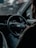 A person is seated in the driver's seat of a car, holding the steering wheel with one hand. The dashboard is visible, showing speedometer dials with faint red accents. The interior appears dimly lit, and there's a sense of focus and concentration.
