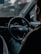 A person is seated in the driver's seat of a car, holding the steering wheel with one hand. The dashboard is visible, showing speedometer dials with faint red accents. The interior appears dimly lit, and there's a sense of focus and concentration.