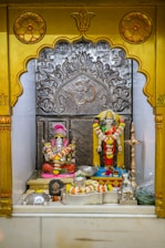 A small altar featuring two Hindu deities adorned with vibrant flower garlands. A statue of Lord Ganesha is seated on the left, with bright red and pink accents. To the right stands a statue of another deity, intricately decorated with yellow and orange floral garlands. There is an ornate silver backdrop with the 'Om' symbol prominently displayed. Various religious items and offerings, such as small vessels and more flowers, are placed in front of the figures.