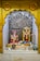A small altar featuring two Hindu deities adorned with vibrant flower garlands. A statue of Lord Ganesha is seated on the left, with bright red and pink accents. To the right stands a statue of another deity, intricately decorated with yellow and orange floral garlands. There is an ornate silver backdrop with the 'Om' symbol prominently displayed. Various religious items and offerings, such as small vessels and more flowers, are placed in front of the figures.