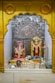 A small altar featuring two Hindu deities adorned with vibrant flower garlands. A statue of Lord Ganesha is seated on the left, with bright red and pink accents. To the right stands a statue of another deity, intricately decorated with yellow and orange floral garlands. There is an ornate silver backdrop with the 'Om' symbol prominently displayed. Various religious items and offerings, such as small vessels and more flowers, are placed in front of the figures.
