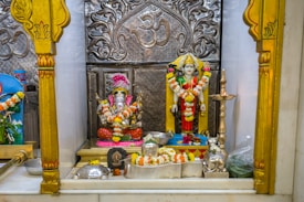 A decorative shrine featuring two adorned statues, one of Lord Ganesha with garlands, and another deity figure next to it. The backdrop is intricately designed with silver engravings and traditional motifs. There are offerings like flowers and small metallic items placed around the statues.