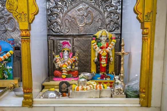 A decorative shrine featuring two adorned statues, one of Lord Ganesha with garlands, and another deity figure next to it. The backdrop is intricately designed with silver engravings and traditional motifs. There are offerings like flowers and small metallic items placed around the statues.