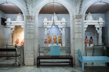 A richly decorated temple interior with ornate white marble arches framing small shrines. Each shrine contains colorful statues adorned with vibrant garments and jewelry. The central aisle has a hanging bell and a seating area with wooden benches.
