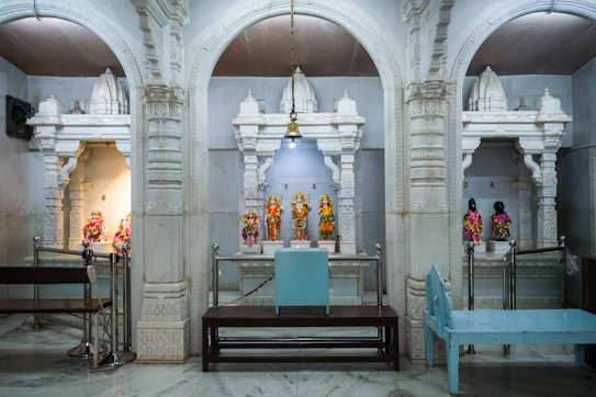 A richly decorated temple interior with ornate white marble arches framing small shrines. Each shrine contains colorful statues adorned with vibrant garments and jewelry. The central aisle has a hanging bell and a seating area with wooden benches.
