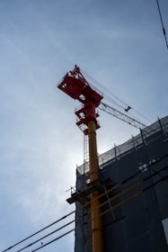 A tall construction crane is positioned next to a building covered in scaffolding. The crane has a bright red upper structure and a yellow shaft, contrasting against a clear blue sky.