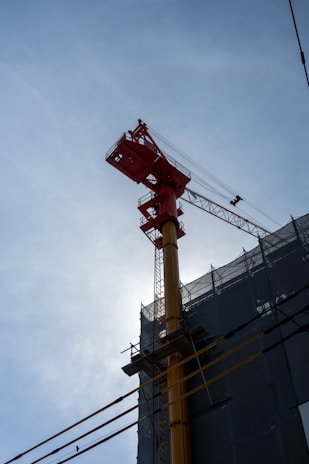 A tall construction crane is positioned next to a building covered in scaffolding. The crane has a bright red upper structure and a yellow shaft, contrasting against a clear blue sky.