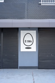 A building facade featuring two closed black shutters and a sign with a black egg shape and the word 'EtWo' on a grey brick wall.