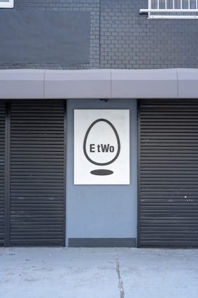 A building facade featuring two closed black shutters and a sign with a black egg shape and the word 'EtWo' on a grey brick wall.