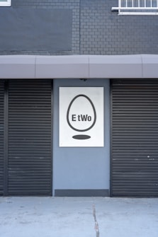A building facade featuring two closed black shutters and a sign with a black egg shape and the word 'EtWo' on a grey brick wall.