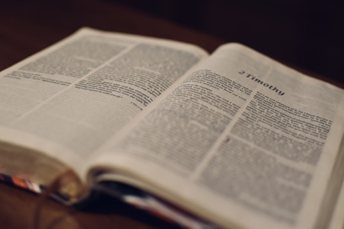 An open book with a page displayed prominently, featuring the heading '2 Timothy'. The text is in a serif font, and the book is positioned on a wooden surface. The pages appear slightly worn, and the focus is on the text.