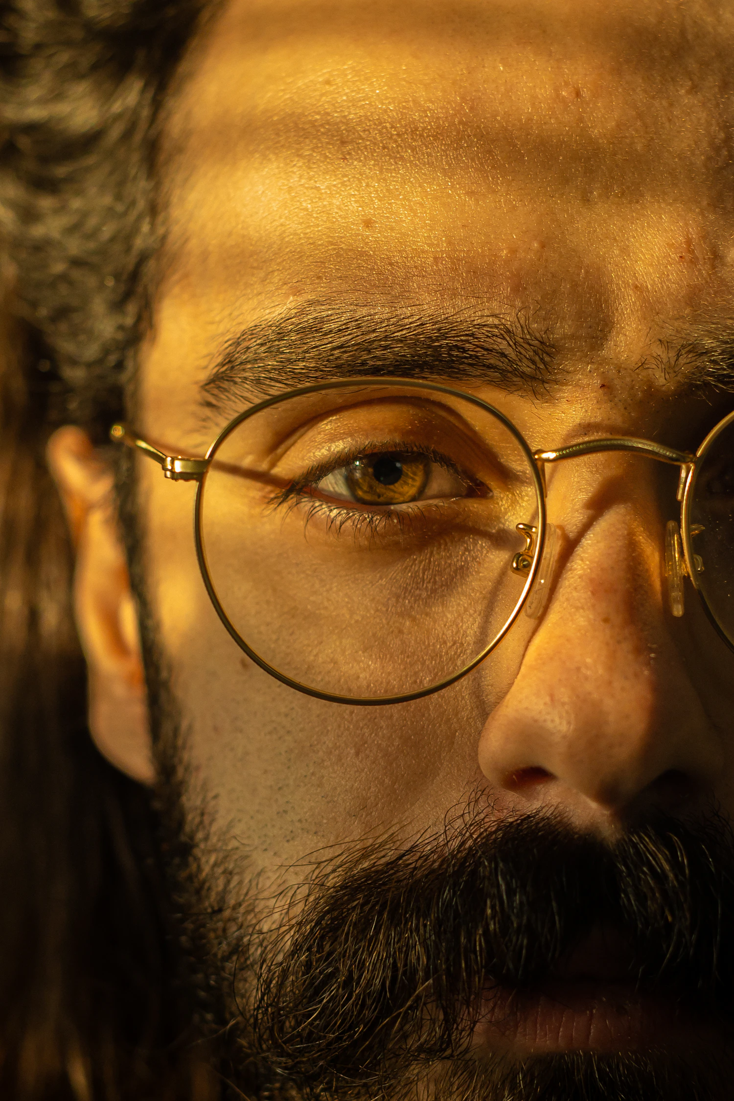 A close view of a bearded man in thin gold round frames under warm low light