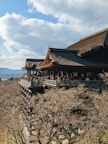 A traditional wooden temple with a large veranda stands amid trees in a hilly landscape. The temple is crowded with people, and the background includes a blue sky with scattered clouds.