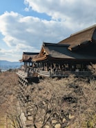 A traditional wooden temple with a large veranda stands amid trees in a hilly landscape. The temple is crowded with people, and the background includes a blue sky with scattered clouds.