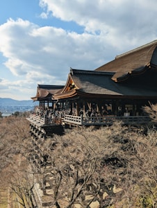 A traditional wooden temple with a large veranda stands amid trees in a hilly landscape. The temple is crowded with people, and the background includes a blue sky with scattered clouds.