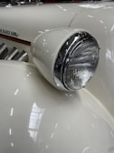 Close-up of a vintage car's front headlight with a white and chrome finish. The car's elegant, curved design is accentuated by smooth lines and a polished surface, highlighting its classic style. Part of the car's emblem and side exhaust pipes are visible, adding to the retro aesthetic.
