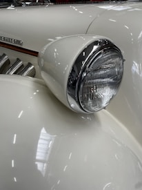 Close-up of a vintage car's front headlight with a white and chrome finish. The car's elegant, curved design is accentuated by smooth lines and a polished surface, highlighting its classic style. Part of the car's emblem and side exhaust pipes are visible, adding to the retro aesthetic.
