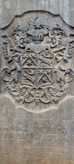An ornate stone carving featuring a coat of arms with intricate scrollwork and heraldic symbols. The shield is divided into sections with various symbols, including a star and animal figures. Above the shield is a knight's helmet with elaborate plumes. Below the coat of arms, there is an inscription in Dutch.
