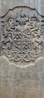 An ornate stone carving featuring a coat of arms with intricate scrollwork and heraldic symbols. The shield is divided into sections with various symbols, including a star and animal figures. Above the shield is a knight's helmet with elaborate plumes. Below the coat of arms, there is an inscription in Dutch.