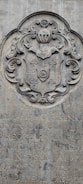 A stone engraving featuring an ornate coat of arms with intricate scrollwork and a shield motif. Below the coat of arms, there is text in a carved script, partially obscured and weathered over time.