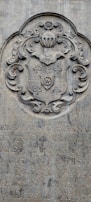 A stone engraving featuring an ornate coat of arms with intricate scrollwork and a shield motif. Below the coat of arms, there is text in a carved script, partially obscured and weathered over time.
