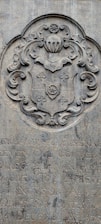 A stone engraving featuring an ornate coat of arms with intricate scrollwork and a shield motif. Below the coat of arms, there is text in a carved script, partially obscured and weathered over time.