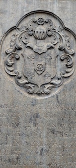 A stone engraving featuring an ornate coat of arms with intricate scrollwork and a shield motif. Below the coat of arms, there is text in a carved script, partially obscured and weathered over time.