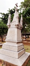 An ornately carved stone monument with a cross on top, surrounded by greenery in a cemetery setting. The monument features detailed architectural elements and a weathered appearance.