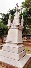 An ornately carved stone monument with a cross on top, surrounded by greenery in a cemetery setting. The monument features detailed architectural elements and a weathered appearance.
