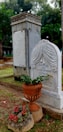 A cemetery scene featuring an ornate tombstone with engraved text and a decorative motif on top. In the foreground, there is a ceramic planter with green foliage and red flowers placed on a stone pedestal. The background includes another taller stone monument and green trees, creating a calm and somber atmosphere.