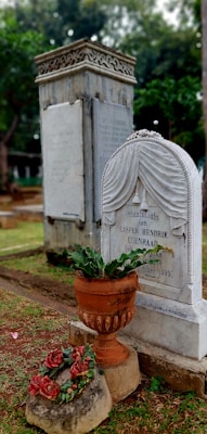 A cemetery scene featuring an ornate tombstone with engraved text and a decorative motif on top. In the foreground, there is a ceramic planter with green foliage and red flowers placed on a stone pedestal. The background includes another taller stone monument and green trees, creating a calm and somber atmosphere.