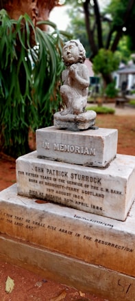 A carved statue of a child is mounted on a gravestone in a cemetery surrounded by trees and greenery. The gravestone features engraved text commemorating John Patrick Sturrock, detailing his years of service and life dates. The atmosphere is somber and tranquil.