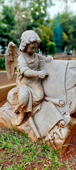 A stone statue of an angelic figure with wings, kneeling and interacting with a large book or scroll. The angel has curly hair and is dressed in flowing robes. It is situated outdoors in a garden or park-like setting with surrounding grass and trees in the background.