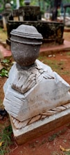 A gravestone in a cemetery features a sculpted urn on top. The stone is weathered with visible dirt and debris around the base. The inscription reads 'Slaap in vrede ontwaak in vreugde.' The surrounding environment includes grass and other tombstones.